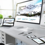 The image depicts a modern office workspace featuring a desktop computer, a laptop, a tablet, and a smartphone, all arranged neatly on a white desk. Each device displays the same content, which is centered around the concept of “RESPONSIVE WEB DESIGN.” The screens show an image of mountains in the background and various browser window sizes, illustrating how the design adapts seamlessly to different screen dimensions. This visual representation highlights the importance of responsive web design, ensuring that web content looks appealing and functions effectively across all devices, from large desktop monitors to small mobile screens. This setup emphasizes the adaptability and consistency of web design in providing a user-friendly experience regardless of the device being used.