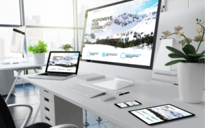 The image depicts a modern office workspace featuring a desktop computer, a laptop, a tablet, and a smartphone, all arranged neatly on a white desk. Each device displays the same content, which is centered around the concept of “RESPONSIVE WEB DESIGN.” The screens show an image of mountains in the background and various browser window sizes, illustrating how the design adapts seamlessly to different screen dimensions. This visual representation highlights the importance of responsive web design, ensuring that web content looks appealing and functions effectively across all devices, from large desktop monitors to small mobile screens. This setup emphasizes the adaptability and consistency of web design in providing a user-friendly experience regardless of the device being used.